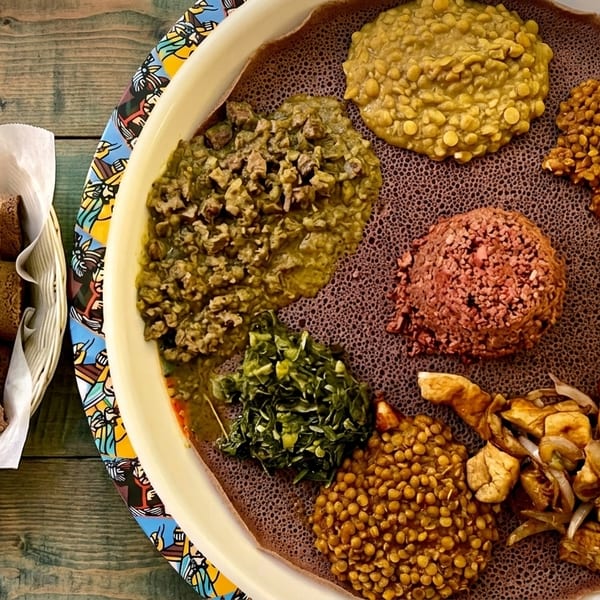 A large platter of Ethiopian injera bread with various colorful stews and a side basket of rolled injera.