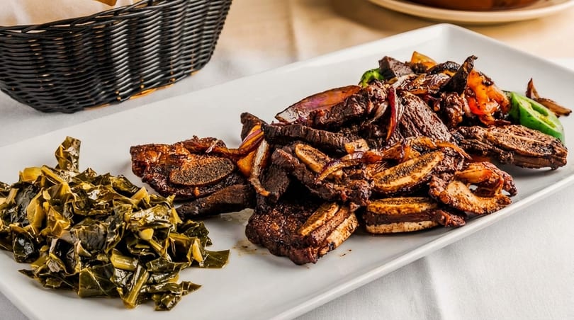 A white rectangular plate holds grilled short ribs, sautéed collard greens, and sliced peppers.