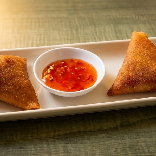 Two golden-brown samosas flank a small white bowl of sweet chili sauce on a rectangular plate.