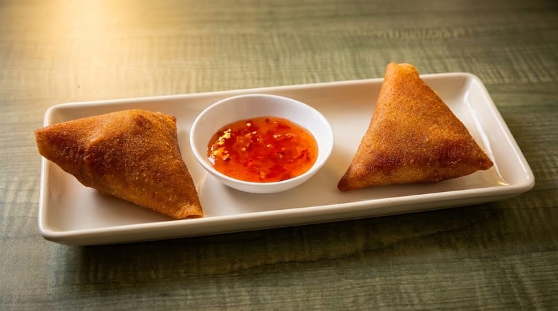 Two golden-brown samosas flank a small white bowl of sweet chili sauce on a rectangular plate.